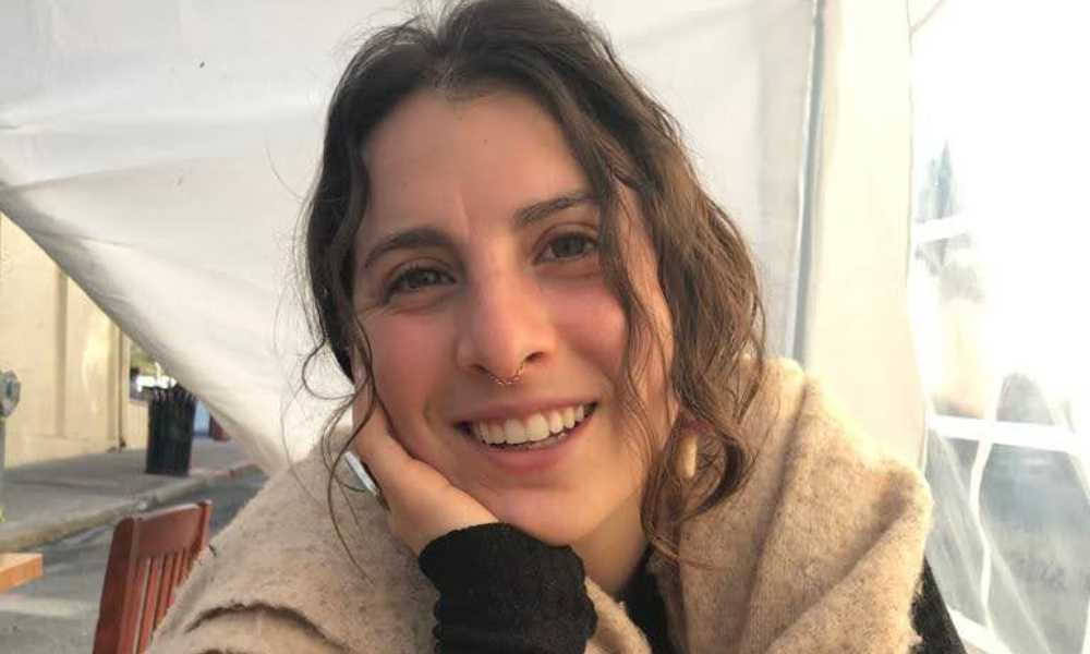 Description: Rachel Binstock, a light skinned brown haired woman, smiles at the camera with her head resting on her right hand.