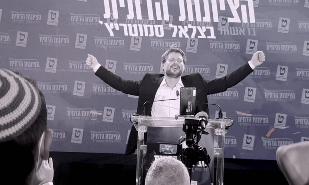 A man in a white collared shirt and a black suitjacket stands at a podium and raises his arms in triumphant celebration.