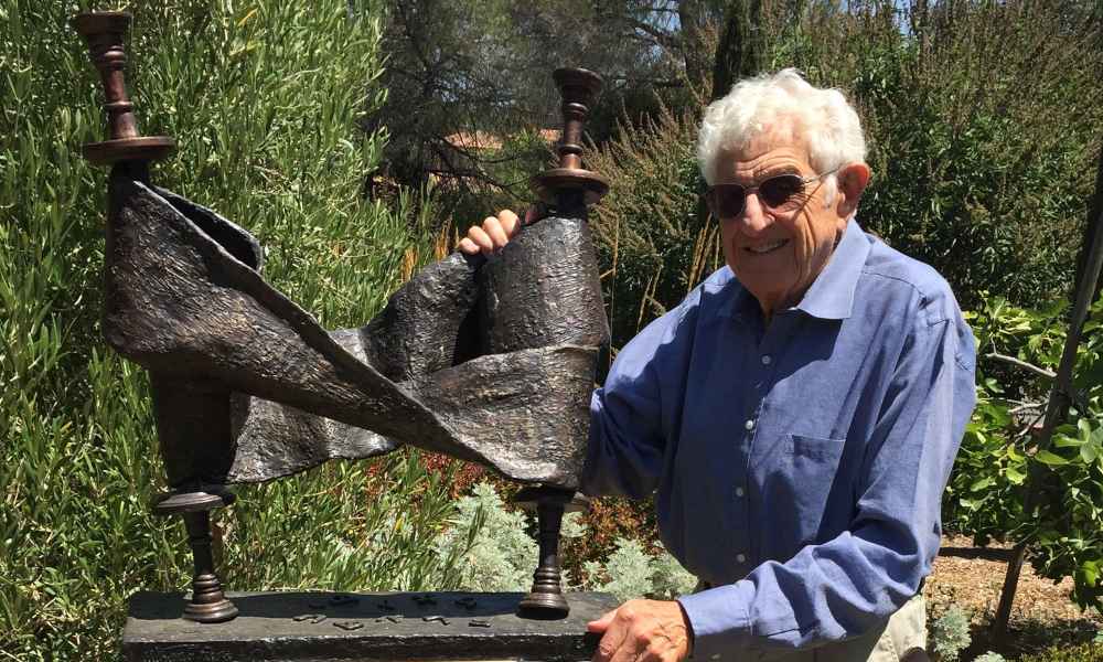 A man with white hair and a blue shirt stands outdoors next to a bronze sculpture.