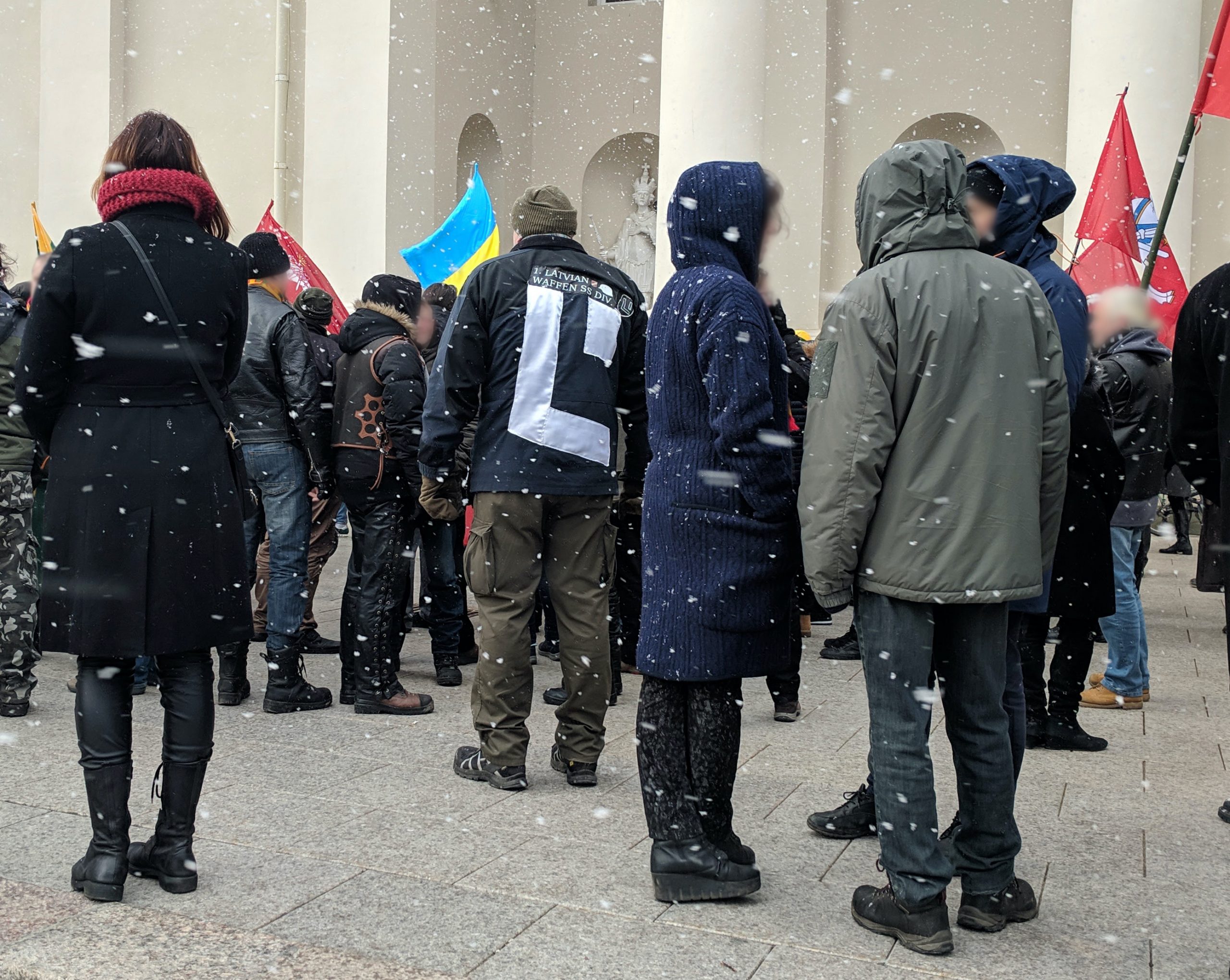 A crowd of people protesting. One man's jacket reads, "Latvian Waffen SS Div."