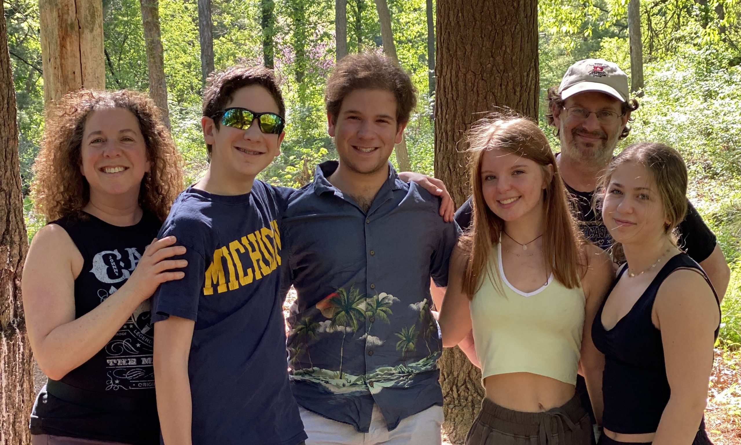 A smiling family stands in the woods. The mother is on the left, the father is second from the right, and the rest are teenagers.