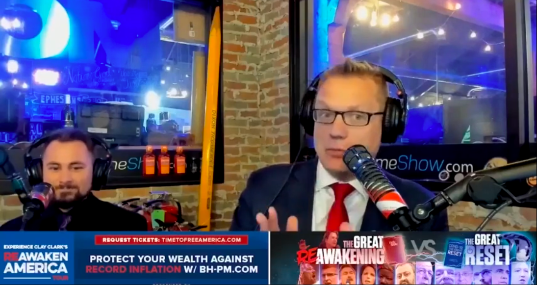 A white man in glasses, a blue blazer, a white shirt, and a red tie speaks into a microphone in a podcast studio.