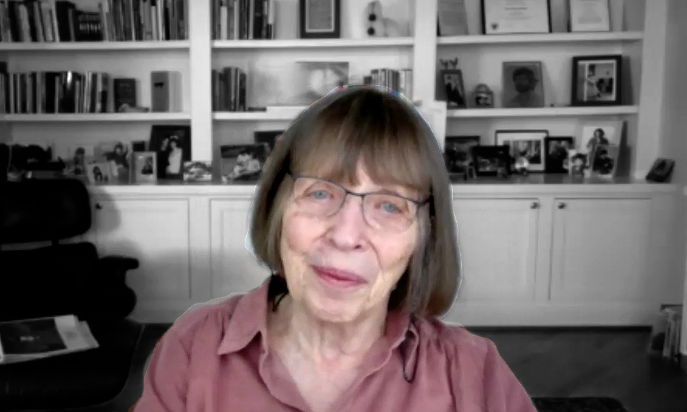 An elderly woman smiles at the camera. She is in color but the background—shelves with books and pictures—is in black and white.