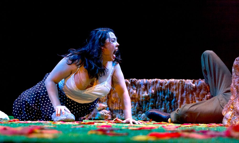 A white woman crouches on the ground while making a ferocious expression. A man's sprawled legs extend from out of the frame.
