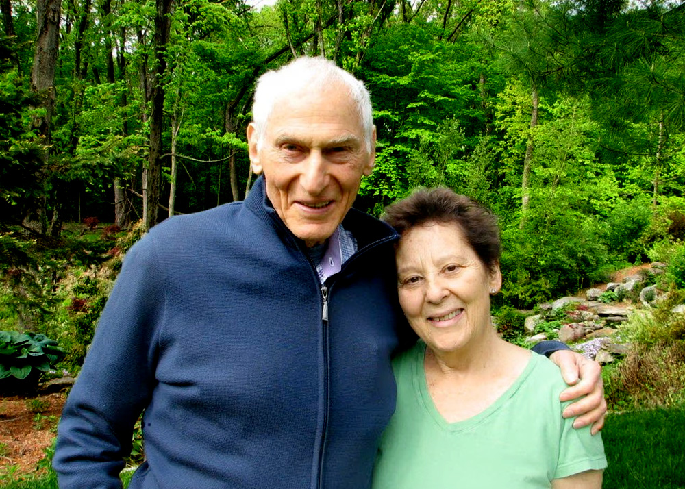 A tall elderly man in a blue cardigan and a woman with brown hair and a green shirt smile at the camera. The man's arm is around the woman's shoulder. Green foliage is behind them.