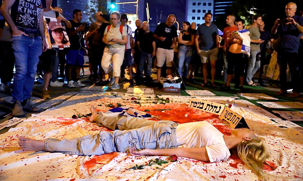 A woman in a white shirt and blue jeans lies still on a white sheet covered with red paint, surrounded by onlookers.