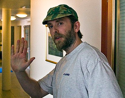 A bearded white man in a grey shirt and camo hat waves at the camera.