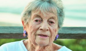 Gloria Levitas—an olive-complexiond elderly woman with short gry hair and purple earrings sits on a bench in front of a large body of water.