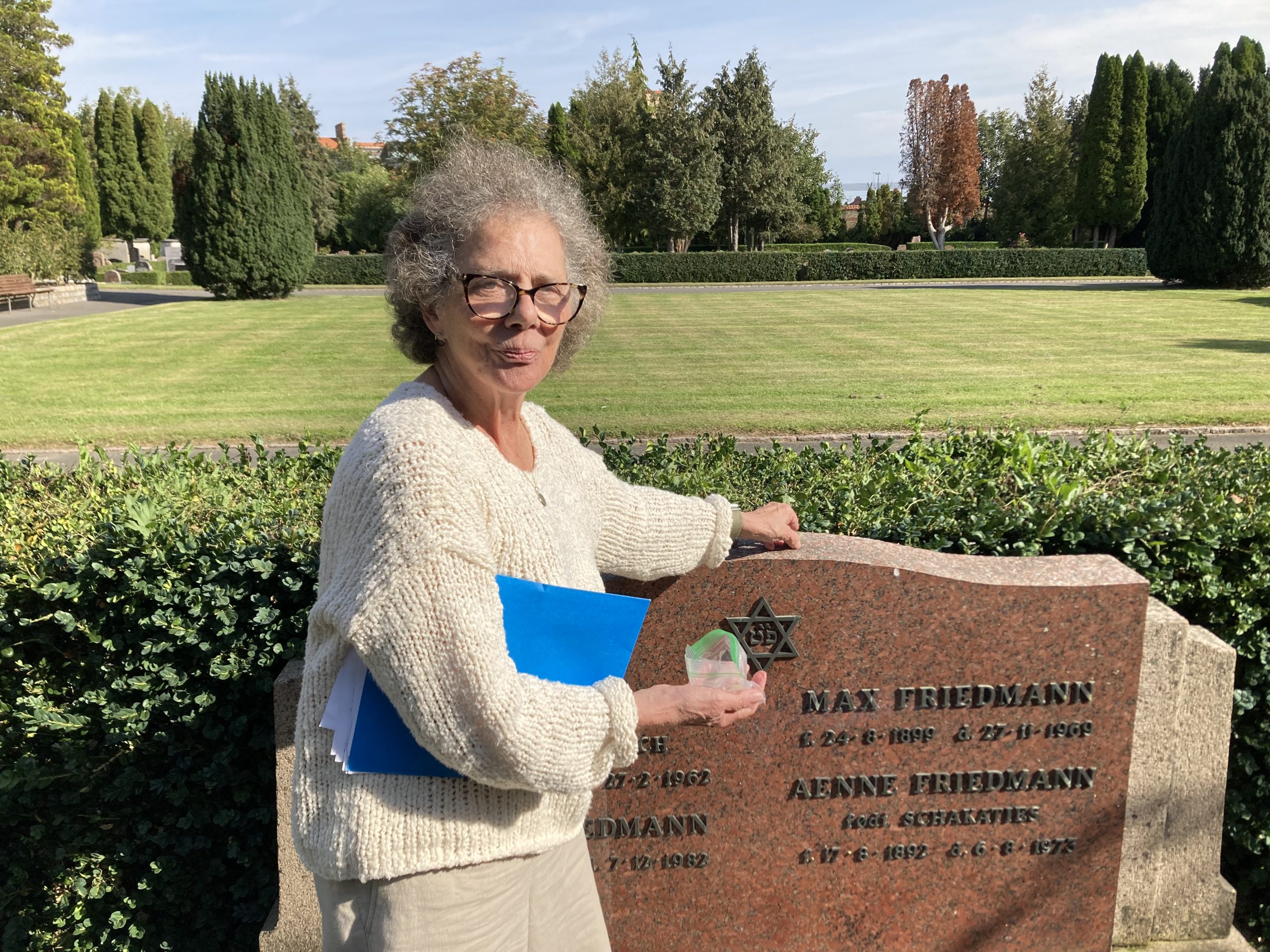 A woman with gray hair with a blue folder under her arm places a stone on a large gravestone.