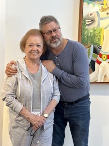 Milch family An older woman in gray smiles. A bearded man hugs her warmly. They are standing in front of a painting.
