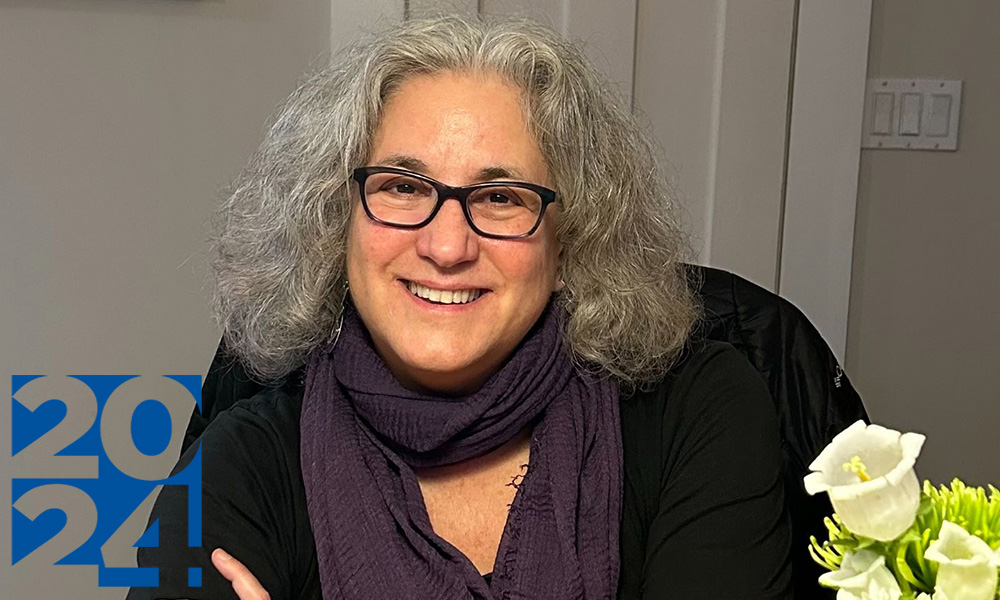 Woman wearing black-framed glasses and a purple scarf; shoulder-length grey hair