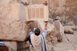 Man wearing kippah, tefillin, and tallit holds up a Torah.