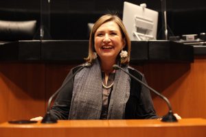 Woman wearing a grey scarf and pearl necklace smiles infront of a microphone.