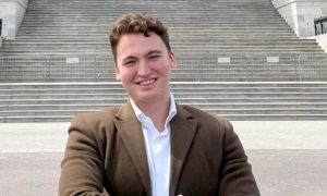 Young man with short, curly hair and wearing a brown blazer smiling.
