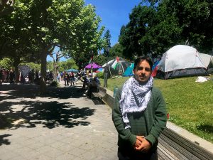 Young man with short dark hair and a mustache, wearing a keffiyeh, stands in front of a line of tents. 
