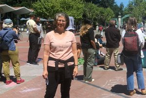 Woman in a peach colored shirt and glasses stands outside among a crowd of people.