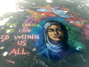 Colorful chalk mural on the ground of a woman in a hijab with an American plane dropping a bomb over her head, the words "The desire to live is with us all" accompany her.