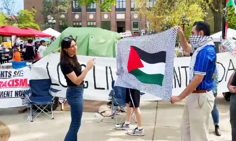 Woman stands outside of pro-Palestine encampment and protesters wave a Palestinian flag in front of her.
