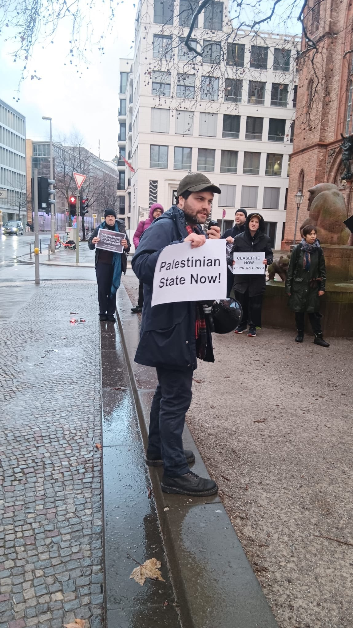 A man holds a sign which says "Palestinian State Now" in black text on white paper. It's a rainy day, and a few other people stand in the background.
