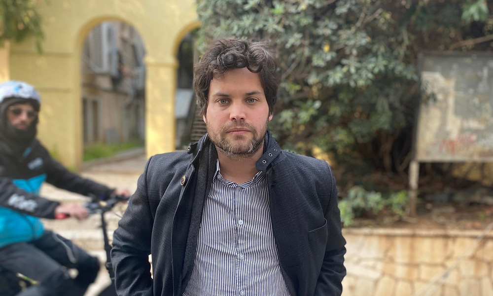 A white man with thick brown hair and a dark jacket looks directly at the camera in front of a quaint urban background. A man in a blue jacket and dark sunglasses wrides a bicycle on the left of the frame and also looks at the camera.