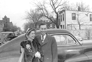 A black and white photo of a smiling couple standing in front of an old car.