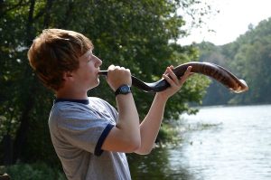 Young man blowing a shofar by a lake.